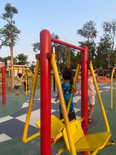 In Sidoarjo, Indonesia, an outdoor playground features children interacting with fitness equipment. A young boy in a light green shirt and blue jeans is seen hanging from a horizontal bar of a red and yellow metal structure. Nearby, another child in a black shirt and grey shorts stands. In the foreground, the focus is on a portion of the red and yellow fitness equipment, with yellow seats visible. The ground is a green surface with a black and white checkered pattern. In the background, trees and other playground structures are present, with a clear sky overhead. The scene appears to be set during daylight hours, likely late afternoon or early evening due to the lighting.