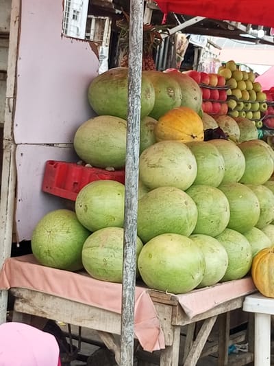 A vibrant outdoor market scene in Pantame, Nigeria, showcases a plentiful display of fresh produce. In the foreground, a collection of large, green watermelons is neatly stacked on a wooden table draped with a pink cloth. Behind the watermelons, a yellow melon, possibly a cantaloupe, adds a contrasting splash of color. Further back, rows of red apples and green pears are meticulously arranged in crates, hinting at a wide variety of fruits available. The market stall is partially covered by a red tarp, providing shade. The background reveals the bustling activity of the market, with glimpses of other stalls and people. The overall impression is one of abundance and lively commerce.