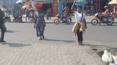 Two women walk along a busy street, each carrying a basket on their head. One woman wears a long blue patterned dress, while the other is dressed in a white shirt, black pants, and a wrapped brown cloth. Motorbikes are parked along the sides of the street and riding down the center. A variety of shops line the street, with people walking in front of them.