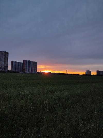 This media file captures a serene sunset scene in Bugry, Russia. The foreground is dominated by a vast, dark green field, likely a meadow or undeveloped land, with tall grasses and wildflowers. In the middle ground, a cluster of large, multi-story buildings, possibly apartment complexes or construction sites, are visible. The buildings are mostly concrete structures, with some appearing unfinished.
The sky above is a spectacle of sunset colors. The horizon is illuminated by a bright orange and yellow glow, indicating the sun is setting or has just set. This warm light contrasts with the cooler, purplish-gray clouds that cover the rest of the sky. The overall atmosphere is peaceful and tranquil, with the fading light casting a soft glow over the landscape.
The time of day is clearly evening, during sunset. The weather appears to be clear, with the exception of the cloud cover. There are no people or animals prominently featured in the image, suggesting a quiet, perhaps even deserted, moment. The location cues, such as the style of the buildings and the presence of a large open field, are consistent with a suburban or semi-rural setting outside of a major city. The image evokes a sense of calm and the quiet beauty of nature at the end of the day, juxtaposed with modern urban development. There is no visible text in the image.