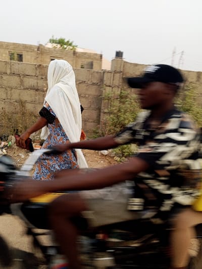 A woman wearing a patterned dress and a white head covering walks beside a man on a motorcycle. The man wears a black baseball cap and a patterned shirt. They are on a street with buildings and vegetation in the background. The image is blurred, suggesting movement. A brick wall is visible behind the woman.