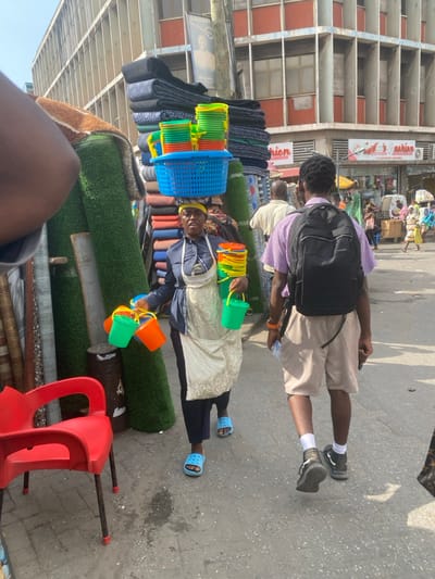 An overhead-angle shot captures a bustling market street in Accra, Ghana, under bright daylight. The primary subject is a dark-skinned adult woman, centrally positioned, walking towards the viewer's left. She wears a dark blue long-sleeved shirt, dark trousers, a light-colored apron, a yellow headwrap, and light blue plastic Crocs. She carries a blue plastic basket filled with stacks of assorted colorful plastic bowls and buckets (yellow, green, orange) balanced on her head. In her left hand, she holds another stack of similar items, and in her right hand, she carries four individual plastic buckets (two green, two orange) by their handles. Her expression is focused.

To her right, a dark-skinned adult male is seen from behind, walking away from the viewer. He wears a striped light purple and white short-sleeved shirt, tan shorts, black sneakers with white socks, and a black backpack. An orange bracelet is visible on his right wrist.

In the foreground, to the left, a red plastic chair with black metal legs is partially visible. Behind it, large rolls of artificial green turf and other rolled materials are vertically stacked, forming part of a market stall. Further back, on the left, more rolled materials are stacked.

The background features a multi-story concrete building with numerous grid-patterned windows. Below it, at street level, are various storefronts, including one with a red sign partially reading "Agatha's Makies" and another visible sign reading "Balian." Numerous indistinct pedestrians and market stalls are visible further down the street. The ground is paved, showing some wear and stains.