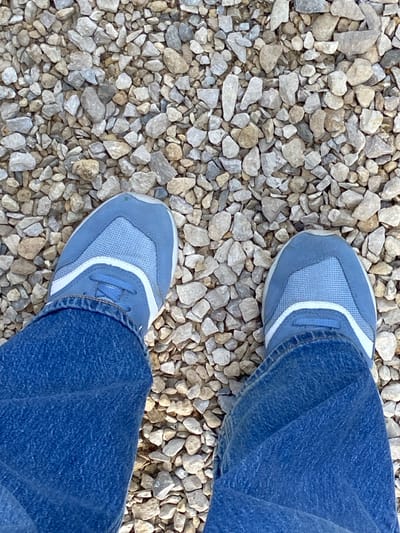 This media file is an overhead shot of two feet wearing blue sneakers and jeans, standing on a surface covered in light-colored gravel. The sneakers are predominantly light blue with white accents. The gravel appears to be a mix of crushed stone and pebbles, typical of an outdoor pathway or ground cover. The image suggests a casual, everyday scene, possibly an individual taking a moment to stand still outdoors. The lighting implies it is daytime, with natural light casting subtle shadows. There are no visible text elements in the image, and no specific location cues beyond the gravel and vegetation which could be found in many outdoor settings. The overall mood is neutral and observational.