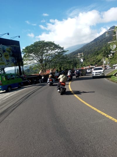 This image captures a bright, sunny day on an asphalt road in Ka Thaung (Upper), Myanmar, traversing a mountainous landscape. The scene features a winding two-lane road with a solid yellow dividing line, where approximately eight individuals on motorbikes are observed traveling away from the camera. The foreground rider wears a light-colored jacket and a helmet. On the left side of the road, a green minivan is parked next to a large billboard displaying advertisements, partially obscured by power lines. Behind these, a large, broad-canopied tree stands amidst roadside structures featuring corrugated roofs. The right side of the road is bordered by a steep, heavily forested mountainside, marked by a series of utility poles. A white SUV is visible further down the road on the right, facing the camera. Clear shadows on the road indicate direct sunlight from a high angle. The sky is bright blue with scattered white cumulus clouds.