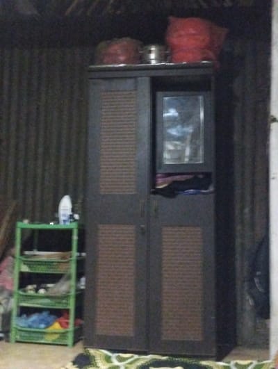 The image captures an indoor scene in Kecamatan Arjasa, Indonesia, characterized by dim lighting. The central focus is a dark brown, two-door wardrobe or cabinet. Its doors feature rectangular panels with a lighter brown, textured or slatted design. An integrated mirror is visible on the upper right door of the cabinet, reflecting a blurry, light-colored area.
On top of the wardrobe, there are several items: two reddish fabric bundles or bags, and a metallic pot positioned between them. To the left of the wardrobe stands a light green, four-tiered plastic shelving unit. The top shelf holds a white bottle, while the lower shelves contain various household items, including what appear to be clothes or other articles stored in baskets.
The background wall is constructed from dark, corrugated metal sheets, suggesting a simple or utilitarian dwelling. A colorful, patterned rug or mat lies on the floor in the foreground. No people are present, and no specific activity is depicted. The subdued lighting indicates the photograph was taken indoors with limited light, possibly in the evening or in an unlit room. No visible text is clearly legible.