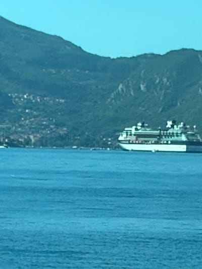 A large cruise ship is anchored in a body of water, with a densely populated hillside and green mountains in the background. The sky is a clear, light blue. The water in the foreground is a deep blue, with subtle ripples. The cruise ship is white and green, with multiple decks and windows. The hillside is covered in buildings, appearing to be a town or village, with trees interspersed. The mountains are a dark green, with some rocky outcrops visible. The image appears to be taken from a distance, possibly from another boat or from the shore.