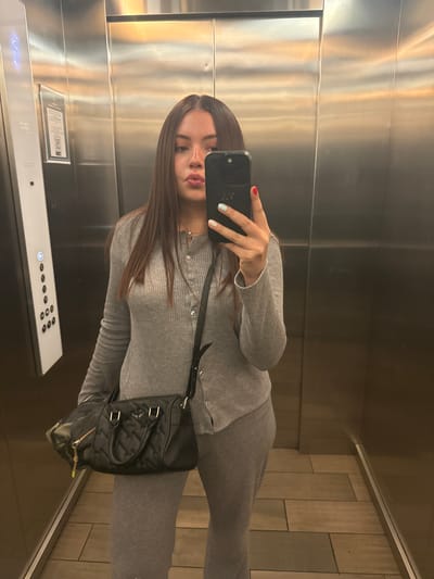 A young woman with long brown hair takes a selfie in an elevator. She is wearing a gray ribbed button-down shirt and matching pants. A black purse is slung across her body. The elevator has stainless steel walls and a panel of buttons. The floor appears to be tiled. The location is inferred to be Mexico City, Mexico.