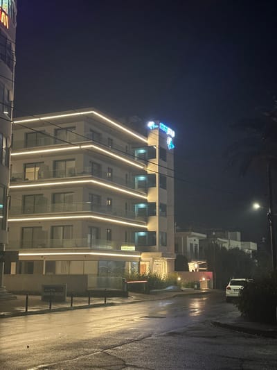 The media file captures a modern, multi-story building illuminated at night, located on a wet street in Egkomi Lefkosias, Cyprus. The building's facade is predominantly white or light-colored, featuring balconies with glass railings and striking architectural strip lighting. Both horizontal and vertical light strips define its structure, casting a warm glow.

At the very top of the building, a prominent blue-teal neon sign reads "THE VIEW". To its right, there appear to be additional characters, possibly in Arabic script, though less clear from this distance. On a yellow sign located on a lower floor, Arabic script is visible, which translates to "View Plaza". Near the bottom right of the image, partially obscured, is another sign that reads "LUXURY APARTMENTS".

The street in front of the building appears wet, reflecting the artificial lights from the building and distant streetlights, suggesting recent rain. A white vehicle, possibly an SUV, is partially visible parked further down the street on the right. A tall palm tree is silhouetted against the dark sky in the upper left, and other scattered greenery is present on the right side of the street and around the adjacent buildings. The overall scene conveys a quiet, modern urban nightscape with no human activity explicitly depicted.