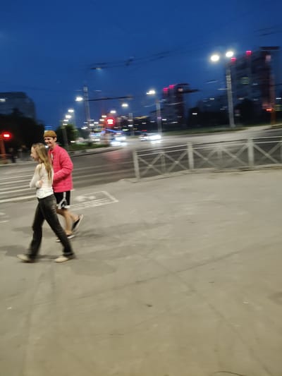 The image shows two individuals walking across a crosswalk at dusk or night in Kemerovo, Russia. The male, on the right, is wearing a bright pink jacket, black shorts, and a brown beanie. He is smiling and looking towards the left. The female, on the left, is wearing a white top and dark pants. She is looking forward and appears to be smiling as well. They are walking in the same direction. In the background, there are city lights, buildings with illuminated windows, streetlights, and traffic on a road. A metal fence is visible in the middle ground. The overall scene suggests a casual urban environment.