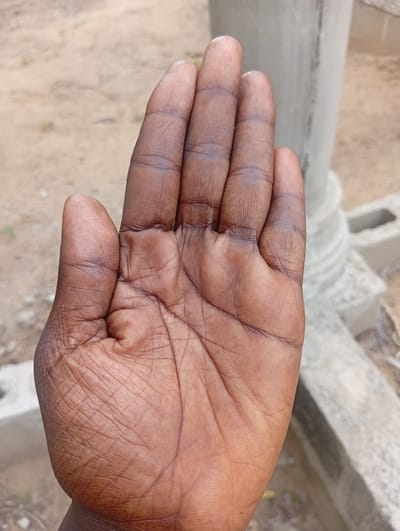 In the city of Gulak, Nigeria, a close-up, eye-level shot focuses on the palm of a person's right hand, held open with fingers extended. The skin is a deep brown, with visible lines and creases characteristic of a palm. The background is blurred, suggesting an outdoor environment with sandy or earthy ground and some construction materials, including concrete blocks and a textured column, likely part of an unfinished building. The lighting appears natural and diffused, creating soft shadows on the hand.