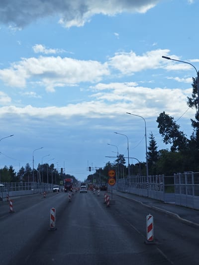 The image depicts a road in Kuznetsovo, Russia, undergoing construction. Several traffic cones and temporary barriers line the road, indicating ongoing work. A red truck and a white car are visible on the road, along with an orange construction vehicle in the background. Overhead, streetlights stand tall, and a bright blue sky with scattered clouds stretches across the frame. The overall scene is one of an active construction zone on a public road.