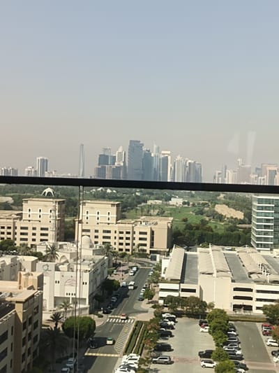 This is a high-angle, medium-shot photograph taken from a building overlooking a cityscape, likely in Dubai, United Arab Emirates, due to the presence of modern architecture and a golf course. The image captures a clear, hazy sky above a dense urban environment filled with various buildings, a golf course, and a street with parked cars.
In the foreground, a railing is visible, suggesting the photo was taken from a balcony or window. Below this, the scene unfolds with multiple buildings of varying heights and architectural styles. Two prominent cream-colored buildings with numerous windows and decorative elements are situated close to each other in the mid-ground. One of these has a minaret-like tower. Further back, a sprawling golf course with lush green fairways and trees stretches across the landscape. Beyond the golf course, a collection of modern skyscrapers dominates the horizon, with some appearing taller and more distinctly designed than others. One particularly striking skyscraper in the distance has a unique, spiraling form.
To the right side of the image, a modern, multi-story white building with a grid-like pattern of windows is visible. Below this, an expansive open-air parking structure with a light-colored canopy covering numerous parking spaces is present. The street level, visible in the lower right and mid-right portions of the image, shows a road with cars parked along the sides and some in motion. Pedestrian crossings are marked on the asphalt. Palm trees and other greenery are interspersed throughout the urban landscape.
The atmosphere appears to be daytime, with soft lighting and a slight haze, which is common in arid environments. The overall impression is one of a modern, developed city with a blend of residential, commercial, and recreational spaces. No people are clearly discernible in the image.