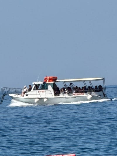 A white tour boat named "POSEIDON" sails on blue water under a clear sky. The boat has a covered seating area where many passengers are visible. The boat is white with red accents on its roof. Two orange life rafts are stacked on the roof of the boat. The boat has a metal ramp at its front. The boat appears to be in motion, creating a wake behind it.