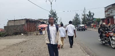 The image displays an urban street scene in Goma, Democratic Republic of the Congo, under an overcast sky. In the foreground, a male individual wearing a white long-sleeved shirt, dark trousers, and a light-colored jacket over a dark backpack walks on an unpaved dirt shoulder, facing slightly right of the viewer. Two additional males walk away from the viewer on the adjacent paved asphalt road. Further down the road, a red motorcycle carrying two individuals, one wearing a patterned helmet/head covering, travels away from the viewer, followed by a dark SUV. The left side of the scene features a partially constructed building composed of concrete blocks with visible rebar, an aged concrete wall, and structures with red corrugated metal siding. The right side of the street is lined with various small commercial buildings, some constructed with corrugated metal, and a red market umbrella with blue plastic chairs is visible near the roadside. Multiple utility poles support an overhead network of wires. Sparse trees are interspersed along the road. The immediate ground on the left is unpaved dirt, while the main thoroughfare is asphalt with a narrow dirt strip on its right edge.
