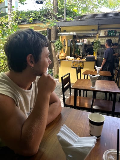 An outdoor seating area at a cafe in Da Nang, Vietnam, features a young man in the foreground, looking to his left with his chin resting on his hand. He wears a light-colored sleeveless shirt. The cafe has a yellow facade with a counter area visible in the background where staff are working. Several wooden tables with black metal frames and chairs are arranged in the seating area. A cup with a straw and a stack of napkins are on a table in the foreground. Lush greenery surrounds the cafe, suggesting a tropical environment.