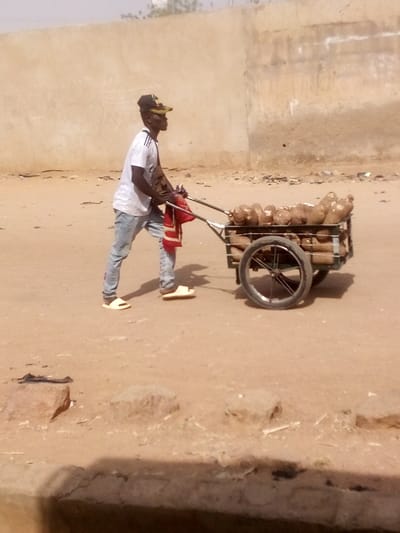 In the city of Tudun Murtala, Nigeria, a lone individual is depicted pushing a two-wheeled utility cart across an unpaved, reddish-brown dirt surface. The person, appearing male, is seen in profile, facing to the right of the frame, actively engaged in pushing the cart forward. He wears a white short-sleeved t-shirt with dark shoulder stripes, light blue distressed jeans, light-colored slide sandals, and a dark baseball cap with a yellow brim. A dark-colored pouch or bag is visible across his right shoulder/chest. His hands grip the metal handles of the cart.
The cart is constructed of dark-colored metal, featuring a rectangular bed positioned over two spoked wheels with dark tires. The cart is heavily laden with numerous large, irregularly shaped tubers, consistent with yams, stacked both within the main cart bed and beneath it. A piece of folded red fabric hangs from the left handle of the cart.
The immediate foreground consists of the same reddish-brown dirt, with scattered small rocks and debris. In the background, a light brown or tan-colored concrete wall or structure with some darker horizontal markings is visible under a hazy sky.