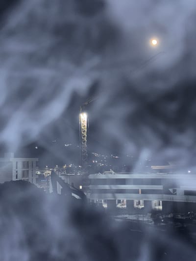 The image depicts a construction site at night in Boreti, Montenegro, enveloped in a heavy mist or fog. A large construction crane, illuminated by bright lights, is the central vertical element of the scene. In the upper right quadrant, a glowing full moon is visible through the atmospheric haze, with what appears to be a faint airplane contrail passing by. In the foreground and midground, various unfinished buildings and construction equipment, including an excavator, are silhouetted against the dark sky and obscured by the mist. The surrounding environment suggests an urban or semi-urban setting with distant city lights visible on a hillside. The overall impression is one of a nocturnal, industrial landscape rendered mysterious by the thick fog.