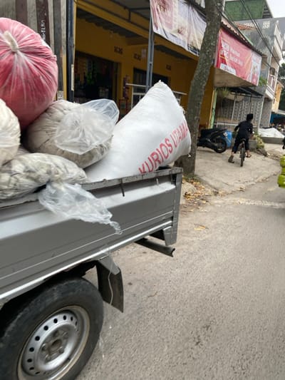 The image shows the back of a silver pickup truck parked on a street in Margaasih, Indonesia, at the coordinates -6.955, 107.55. The truck is loaded with large white bags, a red bag, and white plastic bags. The bags are stacked on the truck bed. The street is paved with asphalt. A man is walking away from the camera on the sidewalk, along a wall with a sign that reads "Makmur Barokah Daya". A tree is in the foreground, and a yellow building is visible in the background. The pickup truck is facing towards the right, and the back tire is visible. The back tire is partially obscured by a large white bag marked with the words "KURIOS UTAMA".