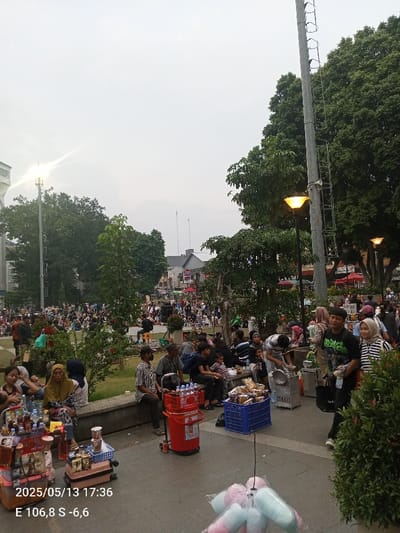 The image shows a street scene in Bogor, Indonesia. The coordinates are: latitude -6.595, longitude 106.792. The scene is a busy street with many people walking and sitting on benches. There is a street vendor selling drinks and snacks. There are many trees and a street lamp in the background. The time is 5:36 PM on May 13, 2023. A street vendor is selling drinks and snacks from a cart. There are people walking past, and many people are sitting and talking on benches. There are a few other street vendors in the background, and many green trees line the street.