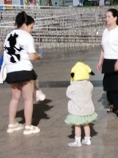 A woman wearing a white t-shirt with a black Mickey Mouse graphic and a black skirt is standing on a paved surface. She has her hair pulled back in a bun. To her right, a young child is dressed in a light gray fuzzy jacket, a light green tutu, and a yellow hat with dark ear-like protrusions. The child is also wearing white socks and sparkly silver shoes. To the right of the child, another woman in a white top and black skirt is visible. In the background, there is a tiered stone structure with railings, suggesting an outdoor seating area or stadium. The lighting suggests it is nighttime or late evening. The overall scene appears to be a casual outdoor gathering.