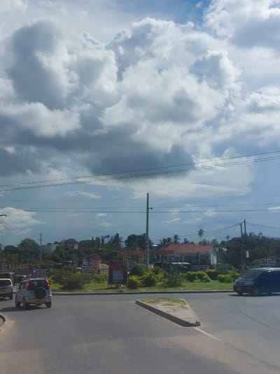 The image presents a daytime exterior view of a multi-lane asphalt road in Dar es-Salaam, Tanzania, beneath a partially cloudy sky with large cumulus formations and visible blue patches. In the foreground, a triangular concrete median with a white painted line separates road lanes. A light-colored compact SUV with a rear-mounted spare tire is observed from its rear-left quarter, moving leftward. On the right side of the road, a dark-colored van is positioned, facing right. No individuals are visible.
The central median is vegetated with green bushes and small trees. Two billboards are located on this median: a prominent one on the right displays the Swahili text "USIKU MWAWA MONDOO," and a smaller, partially obscured sign on the left includes the word "CEMENT." Behind the median, several buildings are visible, notably a structure with a reddish-orange roof and white walls. Overhead power lines and multiple utility poles traverse the scene horizontally and vertically. The background consists of dense tropical foliage, including palm trees, interspersed with additional structures.
