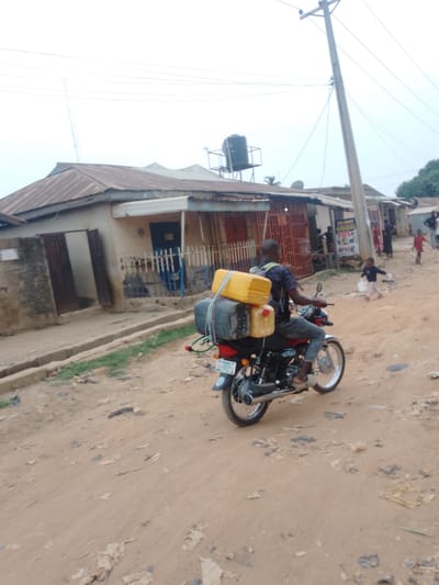 The image shows a man on a motorcycle with two yellow jerrycans and luggage strapped to the back, on a dirt road in Sabon Tasha, Nigeria. The background includes low-rise buildings with rusted metal roofs and deteriorating walls, utility poles, and a water tank on a platform. Several people, including children, are walking near a building with a sign that reads "POS". The road is unpaved and littered with debris.