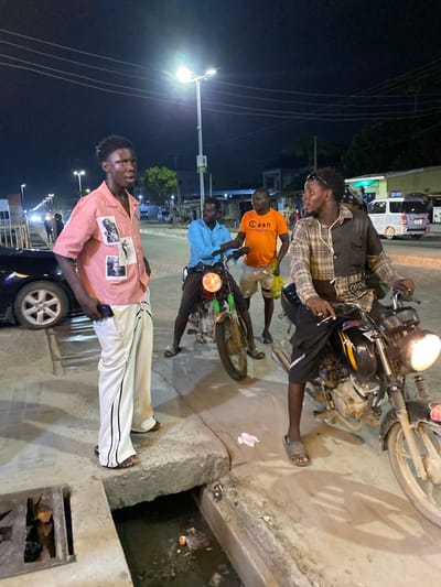 The image depicts a nighttime street scene in Ado, Nigeria. Four men are present, positioned on a sidewalk next to a street. One man is sitting on a motorcycle, wearing a plaid shirt and dark shorts. Another man is wearing an orange t-shirt with the words "Cash Only" printed on it. Another man is wearing a blue shirt. The fourth man wears a pink striped shirt with images on it and white pants. The scene is illuminated by streetlights, with overhead power lines visible. There is another motorcycle adjacent to the one with the man on it. There is a storm drain with visible debris. A parked black car is next to the fourth man. Other vehicles and buildings are also in the background. The image seems to have been taken at night, as indicated by the lighting conditions.