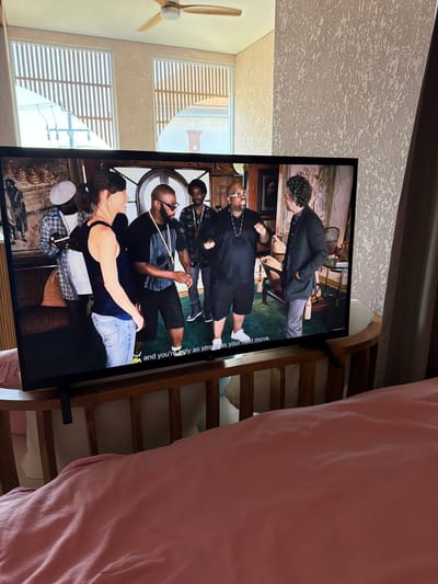 In a bedroom in Mengwi, Indonesia, a flat-screen television displays a scene from a show. The television is set on a wooden stand that is situated in front of a plush pink blanket. The television screen shows a group of five individuals in a room. In the foreground of the television screen is a woman with her back to the camera, wearing a dark tank top and light blue jeans. To her left is a man with glasses and a black shirt, who appears to be speaking. Behind them are three other men, one in a patterned shirt, one in a black shirt with a necklace, and another in a black suit jacket. The room in the television screen has ornate furniture and framed artwork on the walls. The bedroom in the foreground has a ceiling fan and two windows with slatted blinds.