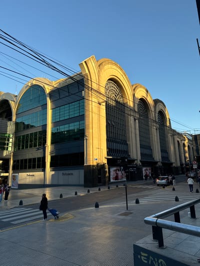 An outdoor, eye-level street view captures a grand building facade and a bustling sidewalk scene, likely in Buenos Aires, Argentina, under a clear blue sky. The building features a distinctive design with several large, prominent arched windows on its upper levels, and modern glass-fronted sections on the lower floors. The architecture, illuminated by warm, golden sunlight from the right, suggests either late afternoon or early morning, casting long shadows across the street.
Numerous black power lines crisscross in front of the building, connecting to poles unseen in the frame. On the ground level, signs for "Parfumerie." are visible on the building's exterior. The street is paved, featuring a zebra crosswalk where a pedestrian in a dark coat walks pushing a blue shopping cart. Other pedestrians are visible further down the street, and a few cars are parked or moving slowly in the background. Street furniture includes bollards lining the pavement and a metal barrier on the far right.
Notable text includes street signs in Spanish. One sign, pointing right, reads "Guardia Vieja" with address numbers "3300 - 3400" below. Another sign, pointing left, indicates "Aguero" with numbers "700 - 800". A 'no parking' symbol is also visible near vehicles in the distance. The overall scene depicts a typical urban environment with a mix of historical architecture and modern elements under bright, clear weather conditions.