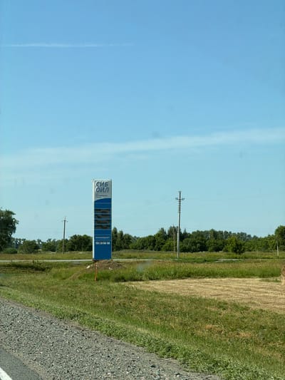 A roadside sign for a "SIB OIL" gas station is prominently displayed in a rural landscape. The sign is blue and white, with the "SIB OIL" logo at the top, followed by text that appears to indicate "service station" in Russian. Below this, there are several rectangular panels that likely display fuel prices, though the numbers are not clearly legible. At the bottom of the sign, it indicates "minimarket" and displays logos for various payment methods, including Visa and Mastercard. The sign is situated in a grassy field with a gravel road in the foreground and trees and a clear blue sky in the background. The environment suggests a sunny day in a location that could be near Bezmenovo, Russia, given the likely Cyrillic script on the sign. There are no people or animals visible in the image.