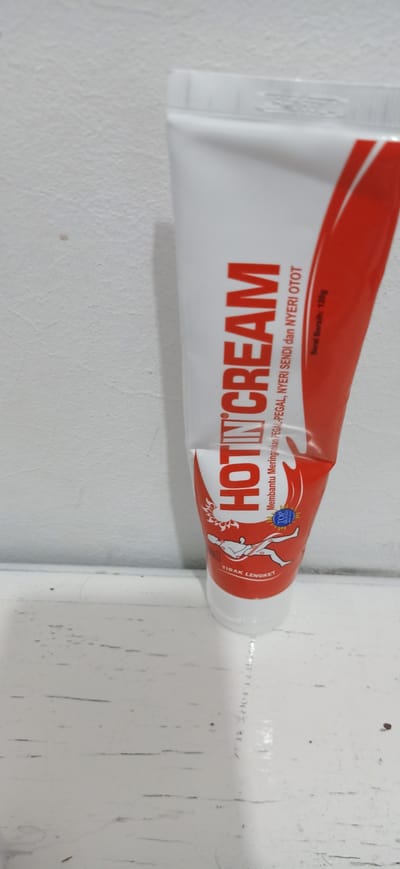 A white tube of "HOTIN CREAM" with red lettering is the primary subject of this medium shot, angled slightly to the right. The cream is designed to relieve muscle aches and joint pain. The tube has a red flame-like design on the right side, and a graphic of a person with lines indicating pain in their legs and back is visible on the lower part of the tube. Text on the tube reads "HOTIN CREAM," "Membantu Meringankan PEGAL-PEGAL, NYERI SENDI dan NYERI OTOT" (which translates to "Helps Relieve Aches, Joint Pain, and Muscle Pain"), and "Berat Bersih: 120g" (Net Weight: 120g). The phrase "TIDAK LENGKET" (Not Sticky) is also visible. The tube is resting on a white surface, possibly a shelf or table, with a white, textured wall in the background. The lighting is bright, suggesting an indoor setting. The overall impression is that of a product shot for a pain relief cream. The location context is Lhokseumawe, Indonesia.