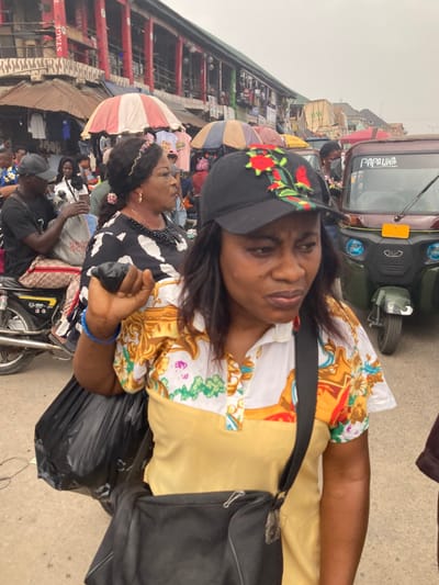 The image depicts a crowded outdoor market street in Aba, Nigeria, under an overcast sky. In the immediate foreground, an adult female of African descent is shown from the chest up, facing forward with a neutral to slightly focused expression. She wears a black baseball cap adorned with red rose embroidery and green leaf accents, a collared short-sleeved shirt with a plain yellow lower section, and a white upper section featuring yellow, orange, and green ornate patterns. A black shoulder bag is worn across her body, and she grasps a large black plastic bag in her right hand, revealing two blue bracelets on her wrist.
To her left, in the midground, an adult female of African descent with dark hair styled with a decorative headband wears a black and white patterned dress, a multi-strand black beaded necklace, and gold hoop earrings. She is looking towards the right. Further left, an adult male, also of African descent, wearing a black baseball cap and t-shirt, sits astride a silver motorcycle, holding a white plastic bag. The motorcycle's seat appears to have a patterned, possibly designer-branded, covering.
The background is densely populated with numerous individuals, market stalls displaying goods, and covered by various umbrellas, including a prominent red and white striped one. A multi-story building with distinctive red vertical structural elements and signage, including "STAGE" and "ARRIA," forms part of the background architecture. A green and black motorized tricycle, labeled "PAPAUWA" on its front, is visible on the right side of the midground. The ground surface is an asphalt or packed earth road, indicating active commerce and pedestrian traffic.