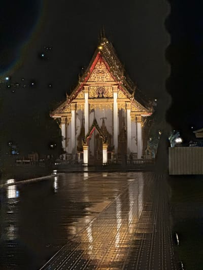 This image captures a nighttime scene in Chalong, Thailand, featuring a temple structure with ornate, golden detailing and a red-tiled roof. The temple appears to be situated on or very near water, as a significant portion of the image is filled with dark water reflecting the temple's lights. The water's surface is disturbed, creating rippling reflections.
To the right of the temple, a long, illuminated structure is visible, likely a pier or a bridge, extending into the darkness. This structure is lined with numerous lights, creating a repeating pattern of illumination and reflection on the water's surface. Some lights are also scattered across the dark water, possibly from boats or other distant sources. A faint rainbow-like arc is visible in the lower left portion of the image, possibly an artifact of the lighting or camera.
The overall mood of the image is serene and slightly mystical, due to the nighttime setting and the interplay of light and shadow on the water. The detailed architecture of the temple suggests it is a significant cultural or religious site. No people are clearly visible in the image. The time of day is clearly night, and the weather appears calm, allowing for the reflections on the water. There is no visible text in the image.