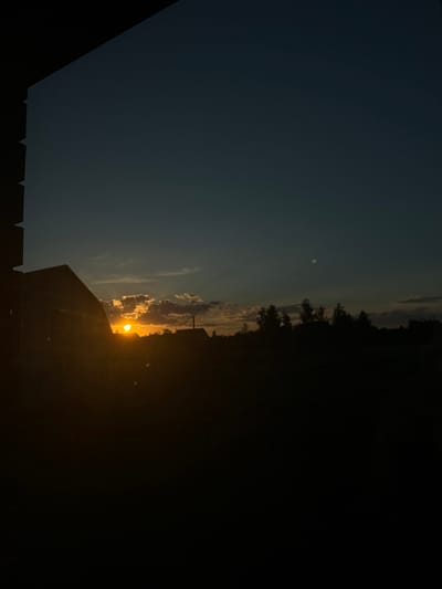 The image captures a sunset over a rural landscape in Chemerne, Ukraine. The sun, a bright orange orb, is setting behind a line of trees, casting a warm glow on the clouds and the sky. The sky transitions from a deep blue at the top to a lighter orange near the horizon. In the foreground, the silhouette of a building and a fence are visible, suggesting a residential area or a farm. The overall mood is serene and peaceful, typical of a sunset. The time of day is clearly evening, and the weather appears clear with scattered clouds. There is no visible text in the image.