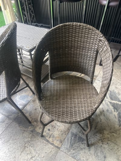 In Howayi, Nigeria, an outdoor patio area features a set of woven wicker furniture. The foreground is dominated by a brown wicker chair with a rounded back and a woven seat. The chair has four legs, with two visible in the image, designed in a slightly curved, ridged style. To the left, partially visible, is another matching wicker chair, and further back, a wicker table with a patterned surface is present. The furniture is situated on a floor paved with large, grey and beige stone tiles. In the background, a dark, slatted metal structure stands, possibly a fence or partition, with hints of greenery beyond. The scene is lit by natural light, suggesting an outdoor setting during the day. There are no people or animals visible in the image.