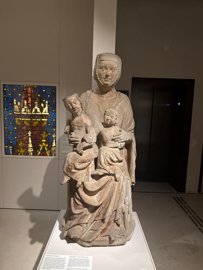 This media file shows a statue of Saint Anne holding the Virgin Mary and the Christ Child. The sculpture is made of stone and appears to be ancient, with worn details and a slightly weathered appearance. Saint Anne is depicted as a mature woman with a serene expression, wearing a headdress and a flowing robe. She cradles the Virgin Mary in her arms, who in turn holds the Christ Child. The figures are rendered with realistic proportions and delicate folds in their garments. The sculpture is displayed in a museum setting, with a muted gray wall and a white pedestal supporting it. In the background, a dark, recessed area suggests a display case or an entrance to another exhibit. Below the sculpture, a framed stained-glass window with a dark blue, starry background and imagery of figures and architectural elements can be seen. There is also a white informational plaque to the right of the sculpture, with text in both German and English, providing details about the artwork. The lighting is artificial and casts subtle shadows, creating a contemplative atmosphere. This artwork is located in Vienna, Austria.