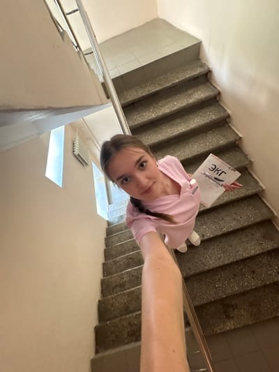 A young woman in a pink scrub suit stands on a concrete staircase, holding a document with Russian text. The staircase has metal railings and is lit by natural light from windows on the wall. The woman is looking at the camera with a slight smile. The background shows a tiled wall and a radiator. The scene appears to be in a public building or institution, possibly a hospital or school, given the attire of the woman and the document she is holding, which seems to be related to medical studies or practice. The specific location in the city of Perm, Russia, is not discernible from the visual information present in the image.