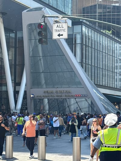 The image depicts a bustling outdoor scene in New York City, likely near Penn Station, as indicated by visible text. A crowd of people, diverse in age and attire, are gathered outdoors. Some are looking towards the camera, while others are engaged in conversation or movement. The architecture suggests a modern urban environment, with a large glass-fronted building dominating the background.
A prominent sign reads "ALL TRAFFIC" with an arrow pointing down, suggesting a road or pedestrian pathway. A traffic light is visible, showing a red signal. The overall atmosphere appears to be that of a busy public space, possibly a transportation hub or a popular gathering spot. The people's expressions are varied, with some appearing relaxed and others more preoccupied. The lighting suggests it's daytime, with sunlight casting shadows, and the weather appears to be clear. The presence of a person wearing a bright yellow safety vest might indicate some form of event management or construction work nearby. The various text elements, including "PENN STATION," "Long Island Rail Road," and subway line identifiers, confirm the location as a major transit point in New York.