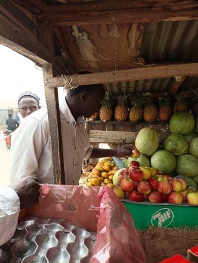 The scene depicts an outdoor fruit stall in Gwagwarwa, Nigeria. The stall is a makeshift structure composed of rough wooden planks and a corrugated metal sheet serving as a roof. A dark-skinned male vendor, wearing a light-colored collared shirt, stands within the stall, reaching with his right hand for a red apple from a prominent pile of red and yellow apples. To the vendor's left, a large mound of yellow-orange mangoes is present. Above and behind the apples and mangoes, a row of eight pineapples hangs from a wooden beam. To the vendor's right, a stack of multiple large green watermelons is visible. In the foreground, the dark-skinned hands of a customer are visible, holding open a translucent red plastic bag inside a gray, molded cardboard egg carton. Behind the vendor, partially obscured by a wooden upright, a second dark-skinned male wearing a dark cap and a light-colored shirt gazes forward. The background shows a dusty, outdoor setting with a blurred street or path, and two distant figures, one on a motorcycle. A green surface bearing a white "7UP" logo is visible beneath the apple display.
