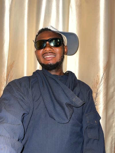 A man in sunglasses and a baseball cap smiles at the camera. He has a dark beard and wears a dark blue collared shirt. The background is a soft, beige fabric with a subtle sheen and a feather design. The image is taken at an angle, with the man positioned sideways relative to the frame.