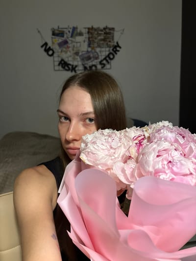 A young woman with long, brown hair and blue eyes is holding a bouquet of pink peonies, partially obscuring her face. She is wearing a black top. In the background, a wire grid is mounted on a light-colored wall, decorated with various pictures and text. The text on the grid reads "NO RISK NO STORY" in black letters. The overall setting appears to be indoors, possibly a bedroom, with a beige chair visible in the foreground. The lighting is dim, suggesting it is nighttime or the room is not well-lit. The specific location within the city of Perm, Russia, cannot be determined from the provided image.
