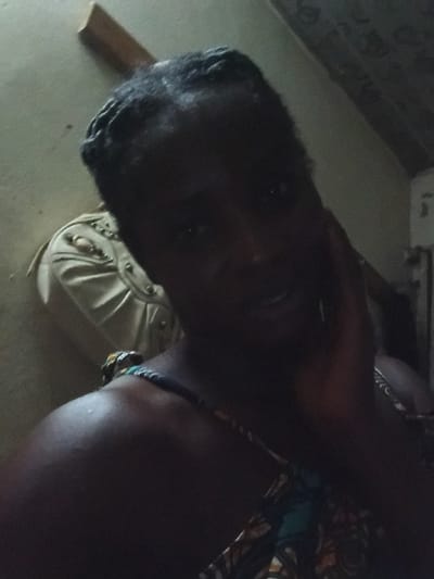 A woman with dark skin, braided hair, and a colorful top rests her head on her hand, looking at the camera. The lighting is dim. Behind her is a cream-colored cushion on a couch, and part of a wall is visible in the background. The image appears to be taken at a tilted angle.
