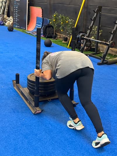 A person is performing a sled push exercise in a gym. The individual, wearing black leggings and a grey t-shirt, is bent over and pushing a weighted sled. The sled has several large weight plates stacked on it. The floor is covered in a bright blue mat. In the background, there is weightlifting equipment, including a rack with a barbell and weights. A small potted plant with yellow fruit is visible to the left of the equipment. There are also black medicine balls on the floor. To the far left, a display screen shows various exercise diagrams and text, some of which is illegible. The lighting suggests an indoor environment, possibly during the daytime.