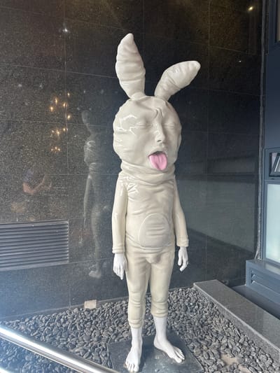 The image shows a sculpture of a reclining human figure with a rabbit head and protruding pink tongue. The figure is pale, glossy, and smooth, with realistic human hands and feet. It wears a tight-fitting garment. The figure lies horizontally on a dark, polished stone floor composed of square tiles. Along the right edge of the image is a border of small dark grey stones. A metal railing is also visible in the lower right corner. The background includes a building facade. The sculpture is located in Madrid, Spain.
