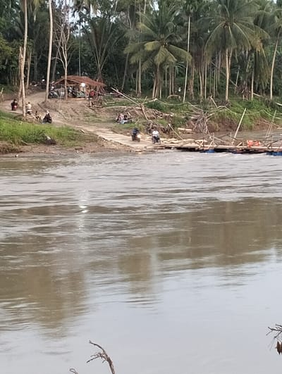 The image shows a wide, muddy river in Kapa, Indonesia. The riverbank is lined with lush, tropical vegetation, including coconut palms and other trees. A small settlement is visible on the opposite bank, with a simple, thatched-roof structure serving as a gathering place. Several individuals on motorcycles are seen approaching or near a makeshift raft bridge across the river. People are also congregating near the riverbank. The river itself reflects the overcast sky, giving it a murky, grayish-brown color.