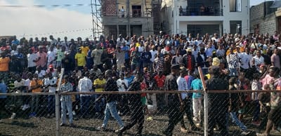 A large crowd of people, mostly men, stand behind a chain-link fence. Some individuals are wearing colorful clothing, while others are in more subdued attire. The crowd appears dense and animated. In the background are partially constructed and finished buildings under a partly cloudy sky. The ground in front of the fence appears to be a dirt surface. The overall scene suggests an event or gathering with significant public interest.