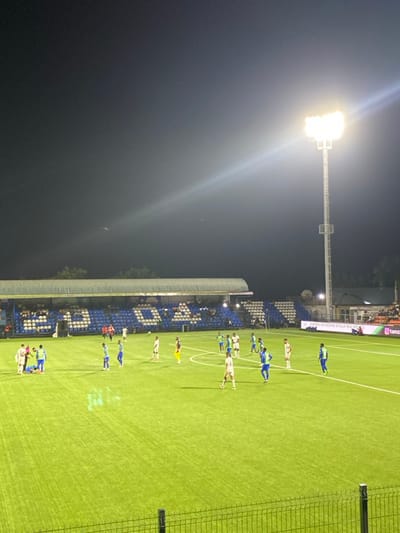 The image shows a soccer field at night, illuminated by a single bright spotlight. The field is green, with white lines marking the pitch. A group of about 10 soccer players wearing blue and white uniforms are standing on the field, seemingly preparing for a match. The players are positioned on the right side of the image, in front of the stadium stands, which are full of spectators. The stands are a vibrant blue color and have a white covering. The image was taken from the stands and the camera is positioned directly behind the players. In the distance, beyond the stands, there are trees and other structures, likely part of the stadium complex. The night sky is dark, with a few stars visible in the background. The location is in Dar es Salaam, Tanzania, at coordinates -6.97 latitude and 39.223 longitude.