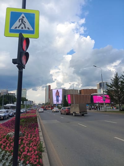 The image depicts a street scene in Perm, Russia, on a partly cloudy day. A pedestrian crossing sign and a traffic light showing red are prominently displayed in the foreground, positioned near a busy road. The road is lined with vibrant pink and purple flowers, creating a decorative median. Several cars and a bus are visible on the road, indicating traffic. In the background, a large modern building with several screens displaying advertisements stands out. One screen features a model in casual clothing, and another shows pink graphics with text. The architecture of the building suggests it is a shopping center or entertainment complex. People can be seen walking along the sidewalk in front of the building. The sky is a mix of blue and dramatic grey clouds, hinting at changing weather. The overall impression is an urban environment with commercial activity and traffic flow.