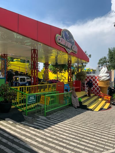 The image shows a brightly colored outdoor area in the city of Batu, Indonesia, located at the coordinates -7.884 latitude and 112.525 longitude. It is a "Convoy Car" ride for children at a local amusement park. The ride consists of small cars that travel on a track. The cars are lined up in front of a red and yellow building with a sign that says "CONVOY CAR" in white letters. Two adults are waiting for the ride to start. One is wearing a black hijab and the other is wearing a white hijab. A child wearing a pink shirt is standing on the stairs waiting for the ride to start. There are two yellow signs with black letters on them that read "MASUK ANTRIAN CONVOY CAR" and "MOHON SEDIKIT PERHATIAN BAWAAN TIDAK BOLEH DI TARUH DI ATAS JALAN, UNDER THE RIDE."  The ground in front of the ride is made of a patterned stone. There are trees and bushes behind the ride.