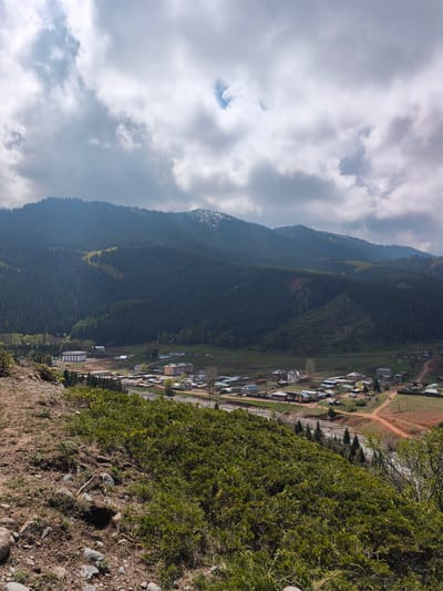 This elevated view captures a serene mountain valley landscape, likely in Jeti-Ögüz, Kyrgyzstan, under a largely overcast sky with patches of brighter cloud and hints of blue. Majestic, dark green, coniferous-covered mountains dominate the background, their slopes rising sharply, with some higher peaks still showing remnants of snow. Portions of the mountains reveal reddish-brown soil.
Nestled in the valley below is a scattered village, characterized by numerous houses and buildings. These structures vary in style, from more traditional, simpler homes to a few larger, more modern-looking buildings, including one appearing to be under construction with a distinctive striped facade. Dirt roads, also reddish-brown in color, crisscross the village and surrounding fields. A river or stream, with a rocky bed, flows through the valley, partly obscured by vegetation and structures.
The foreground consists of a steep, grassy hillside, dotted with rocks and patches of shrubbery in various shades of green and brown. No people or visible text are discernible in the image. The time of day appears to be daytime, possibly late morning or afternoon, given the overall brightness despite the heavy cloud cover. The scene evokes a sense of natural beauty and rural life in a mountainous region.